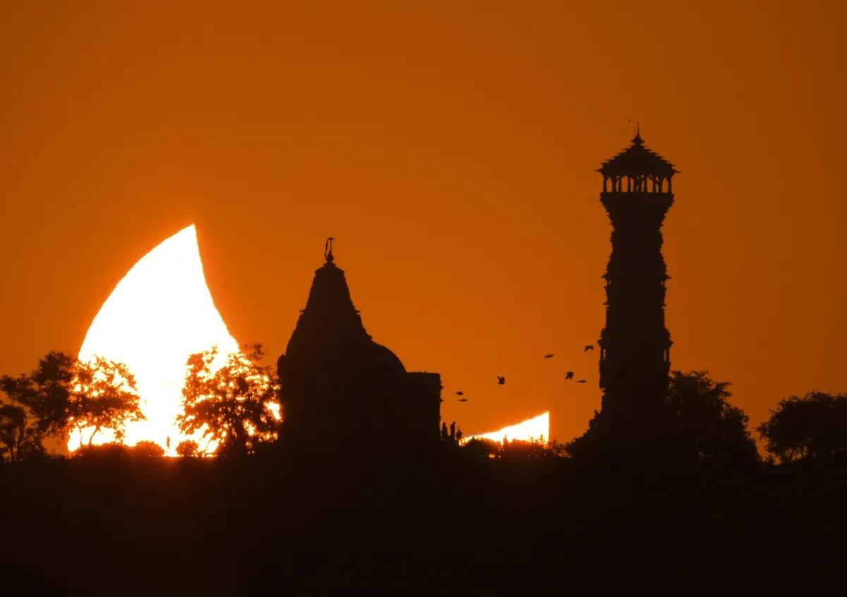 A dramatic landscape photograph of the Sun during a partial solar eclipse. The silhouette of a temple structure is in the foreground, and behind the Sun has been partially obscured by the Moon's shadow.